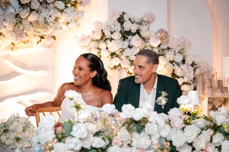 A bride and groom smiling at their wedding reception, surrounded by white and pink floral arrangements and gold accents.