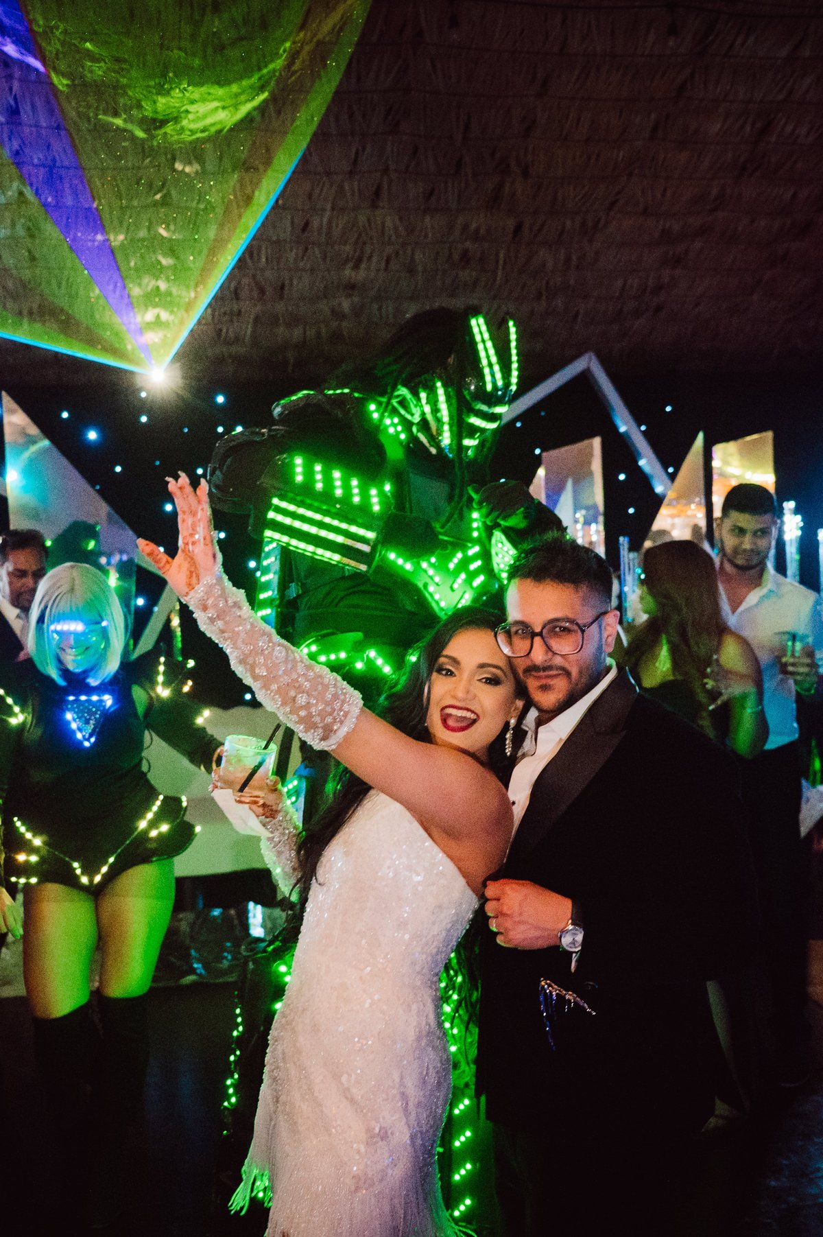 Couple in formal attire posing at neon-lit nightclub event with glowing green geometric structures and crowd in background