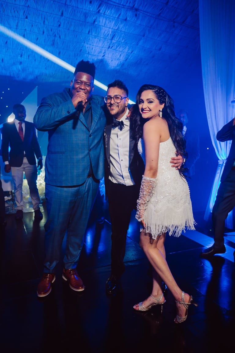 Three people at a formal event with blue stage lighting. Man holding microphone in blue suit, man in tuxedo, and woman in white sequined dress.