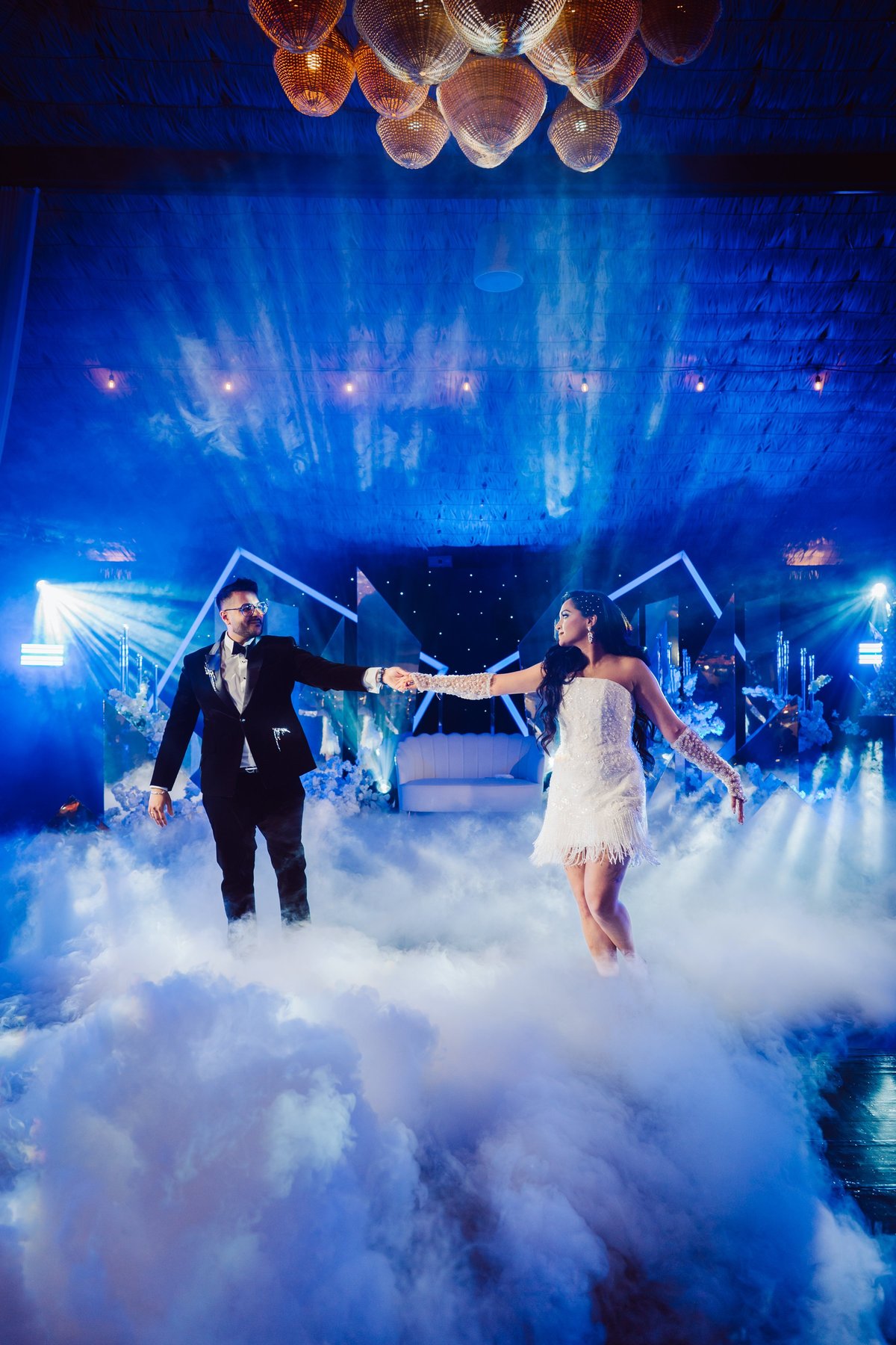 Bride and groom dancing on stage surrounded by dry ice fog with blue lighting and decorative hanging lights above