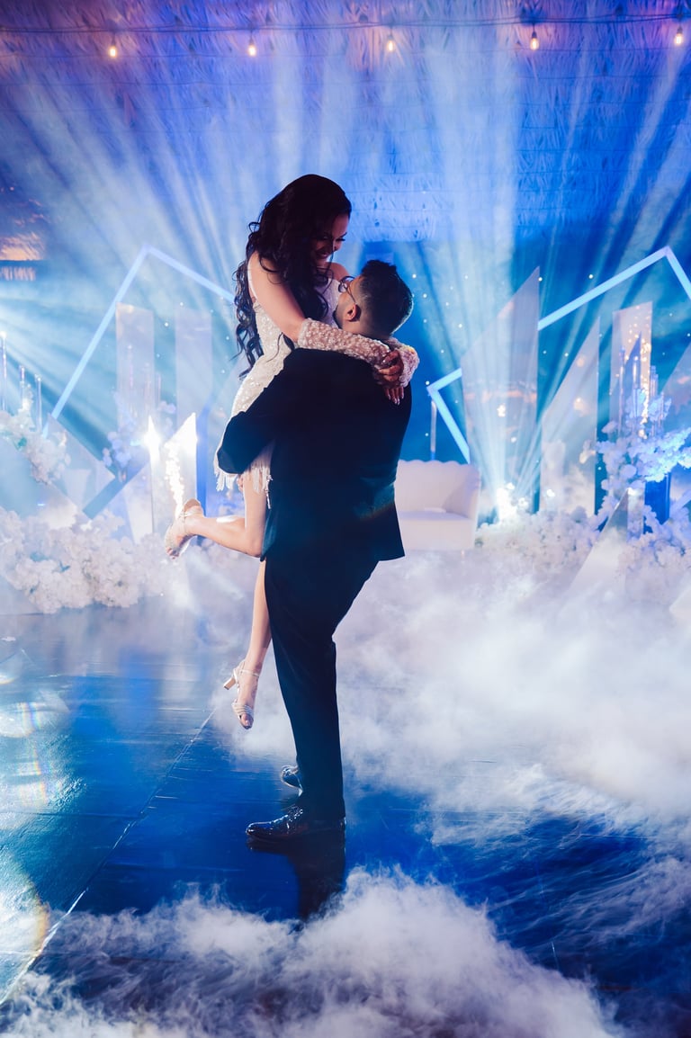 Groom lifting bride in a romantic dance surrounded by blue stage lighting, dry ice fog, and neon geometric shapes