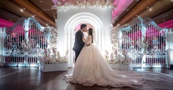 A bride and groom kiss under an illuminated white archway decorated with flowers in an elegant ballroom setting.