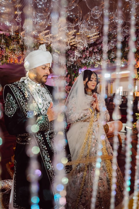 Bride and groom in traditional South Asian wedding attire with ornate embroidery and beaded decorations at an illuminated venue