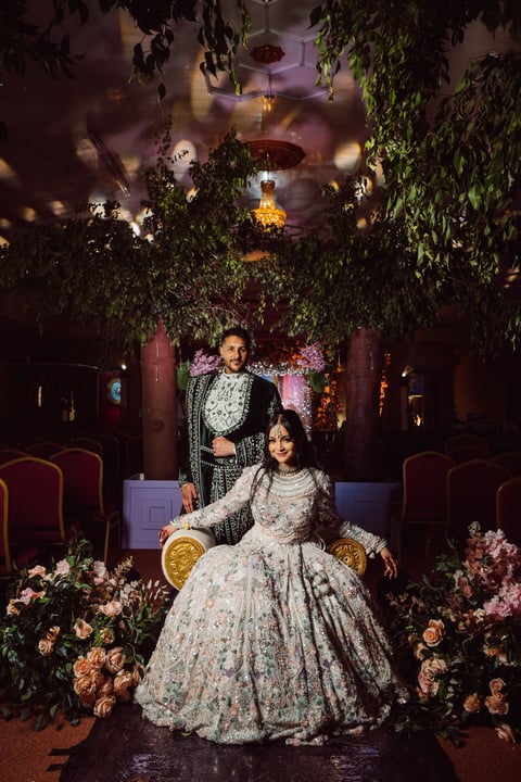 Couple in ornate wedding attire posing in lavishly decorated venue with hanging greenery, floral arrangements, and ambient lighting