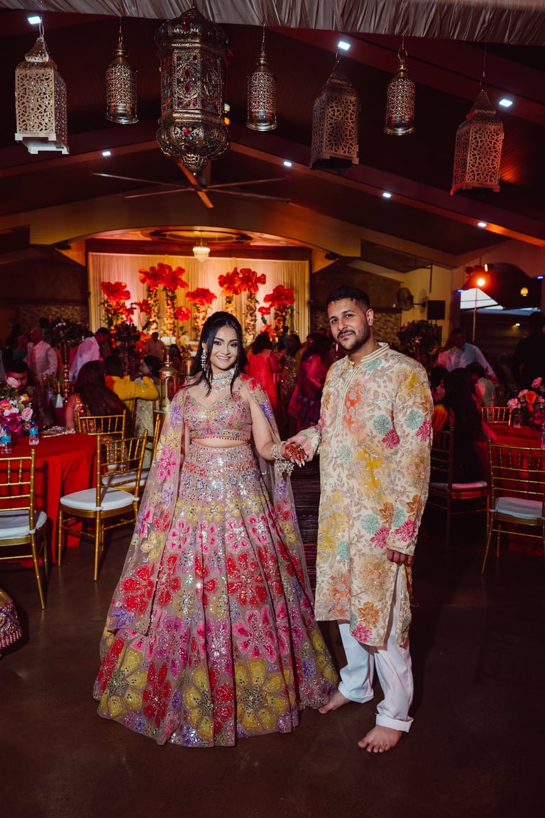 Couple in traditional Indian wedding attire posing in ornate banquet hall with hanging lanterns and floral decorations
