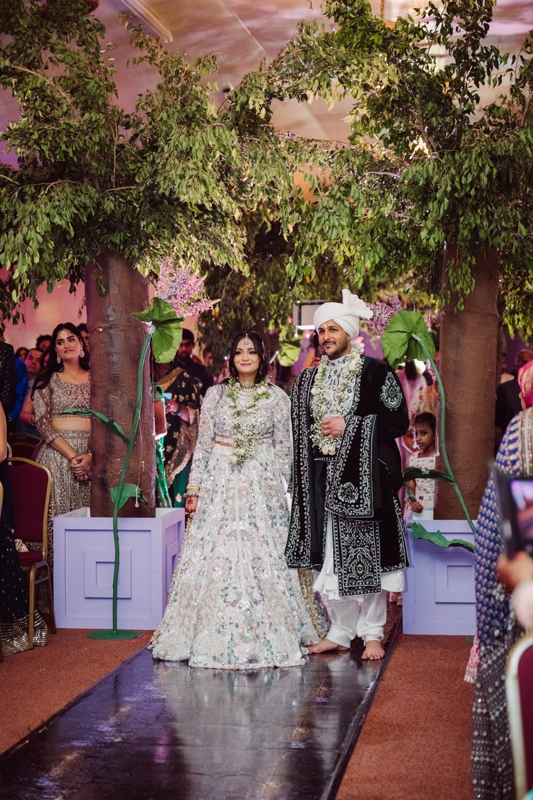 Bride in white embroidered lehenga and groom in black sherwani walking on runway at indoor Indian wedding reception decorated with hanging vines and pink flowers