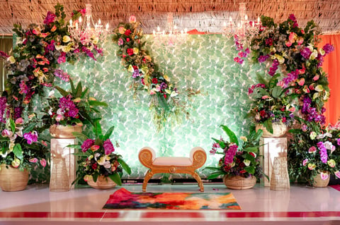 Ornate event space with wooden bench, vibrant pink flowers and greenery arrangements, green sequined backdrop, and colorful rug