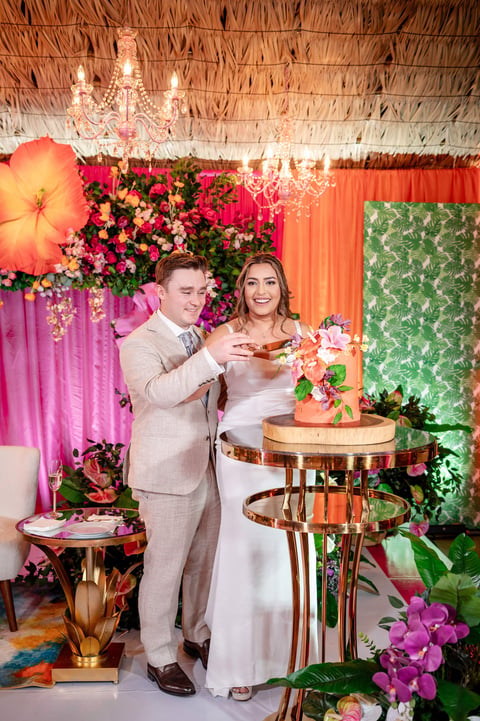 Bride and groom cutting cake at an ornate wedding reception with floral backdrop and crystal chandelier