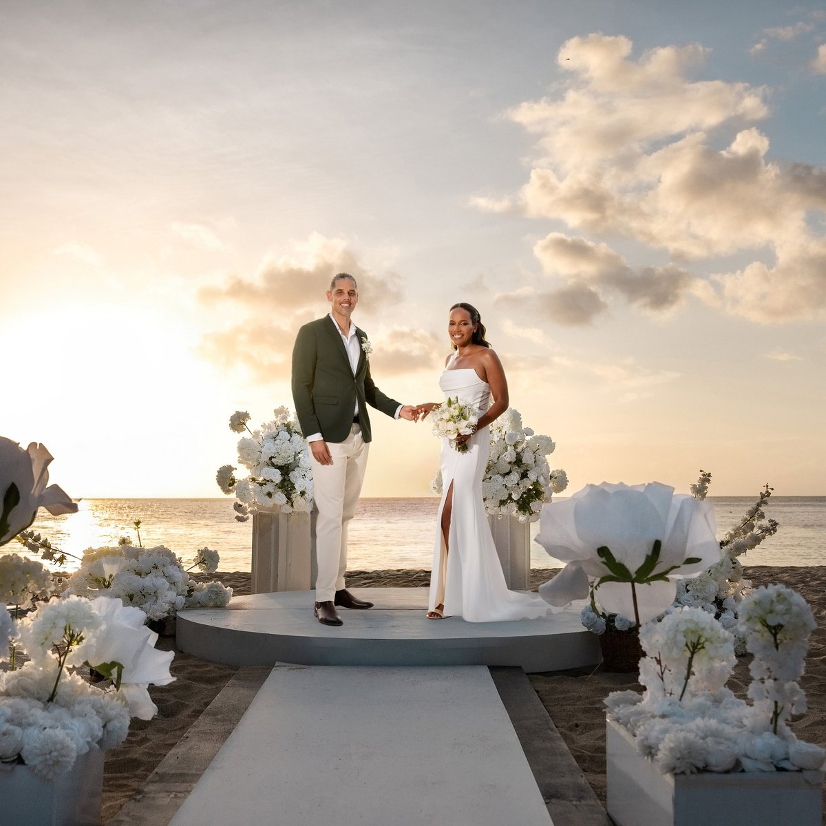 Bride and groom standing on stage during beachfront wedding ceremony with white floral decorations and ocean sunset backdrop