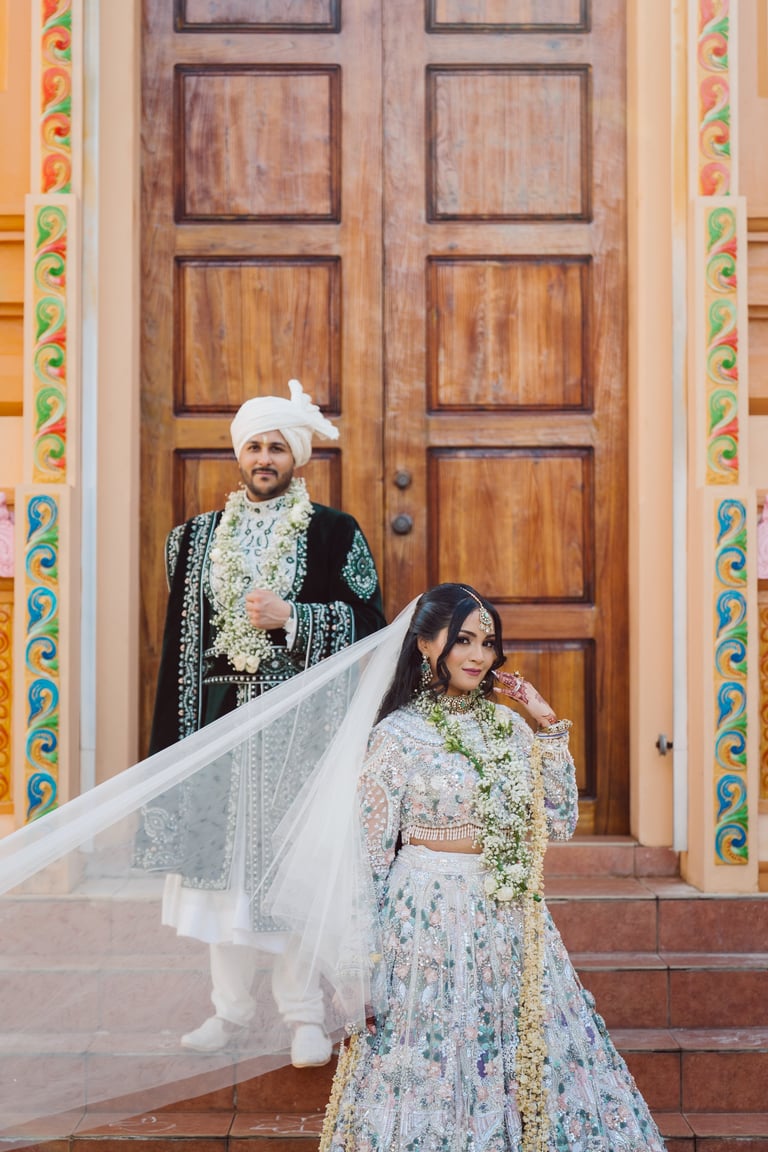 Bride and groom in traditional South Asian wedding attire posing before ornate wooden doors with decorative pillar frames