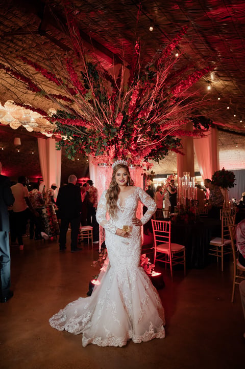 Bride in white lace wedding dress posing in elegant red and gold decorated ballroom with dramatic floral installation