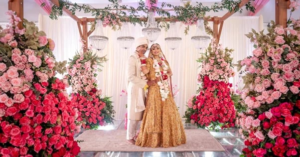 Bride and groom in traditional Indian wedding attire standing beneath a floral arch with pink and red roses
