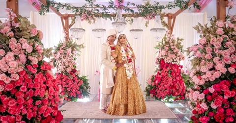 Bride and groom in traditional Indian wedding attire standing beneath a floral arch with pink and red roses