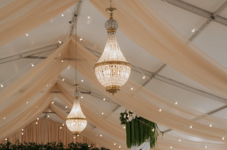 Elegant crystal chandeliers hang from a white tent ceiling with draped fabric and warm string lights