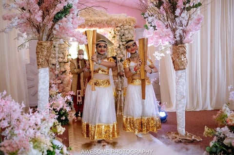 Bride with bridesmaids in white and gold dresses standing in ornate pink and gold wedding ceremony archway with floral decorations