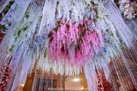 Elaborate floral ceiling installation with cascading white, pink, and purple flowers hanging from strings above a golden light