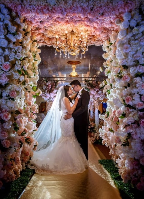Bride and groom kiss under a luxurious floral archway of pink and white roses at their wedding ceremony.