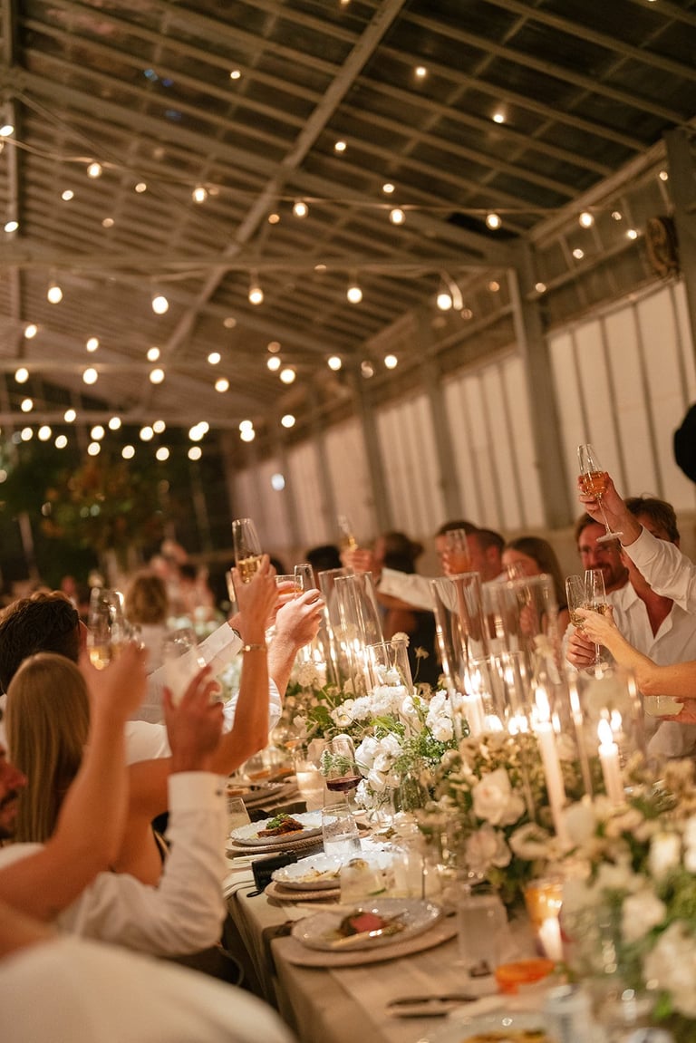 Elegant indoor wedding reception with guests toasting at a long table decorated with white flowers and candles under twinkling string lights