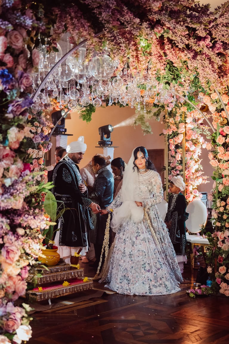 Bride in ornate white wedding lehenga stands under elaborate floral archway with family members at wedding celebration