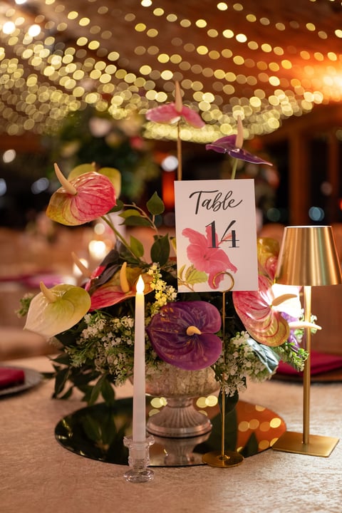 Elegant wedding table centerpiece with Table 14 card, pink and purple flowers, candle, and gold lamp on bokeh lit background