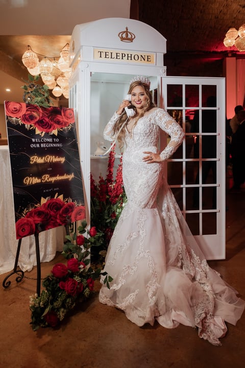 Bride in elegant white lace gown posing beside a vintage white telephone booth with red roses and welcome sign at wedding reception