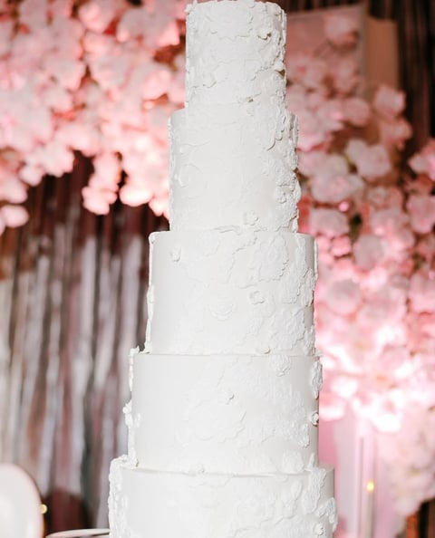 Five-tiered white wedding cake with textured frosting displayed against a backdrop of pink floral arrangements