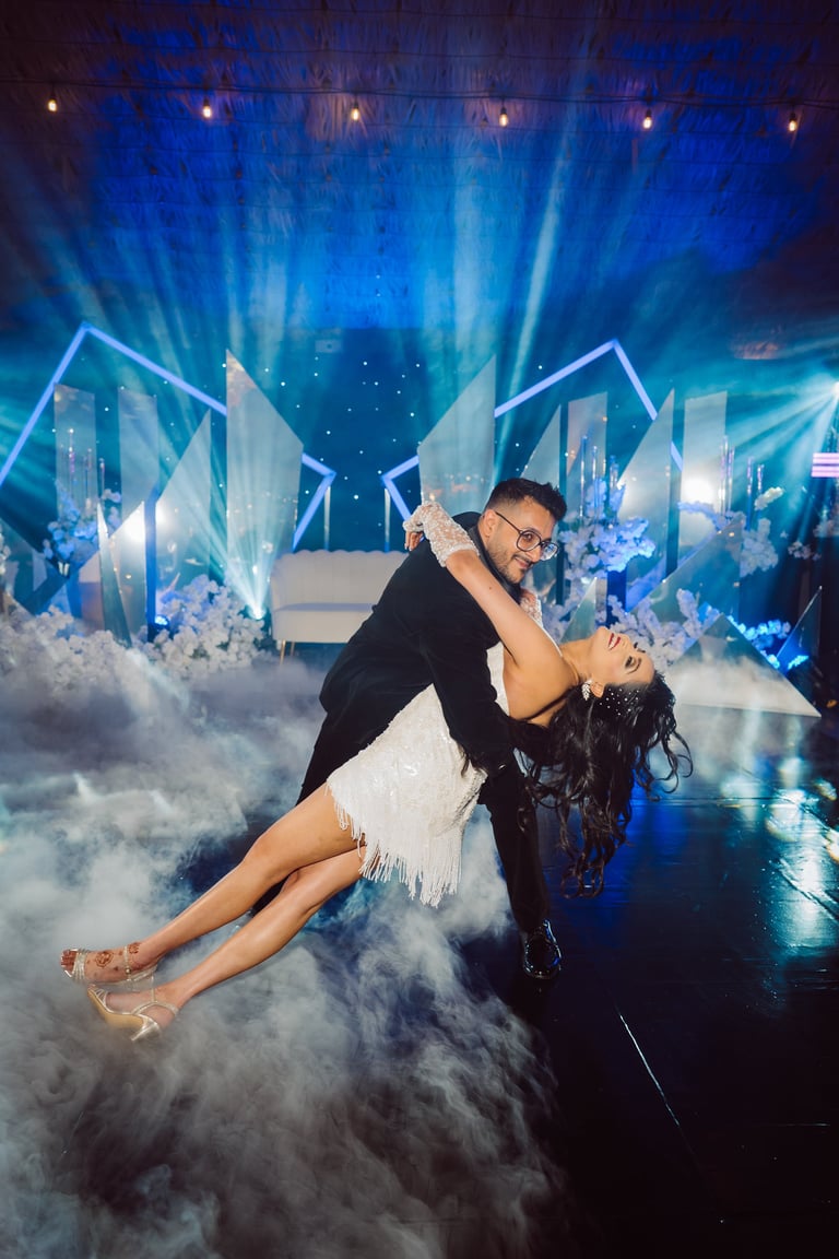 Man in black jacket and woman in white dress performing a dramatic dip dance move on stage with blue lights and fog effects