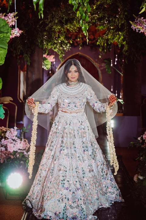 Bride in ornate white embellished lehenga and veil walking down floral-decorated aisle at night