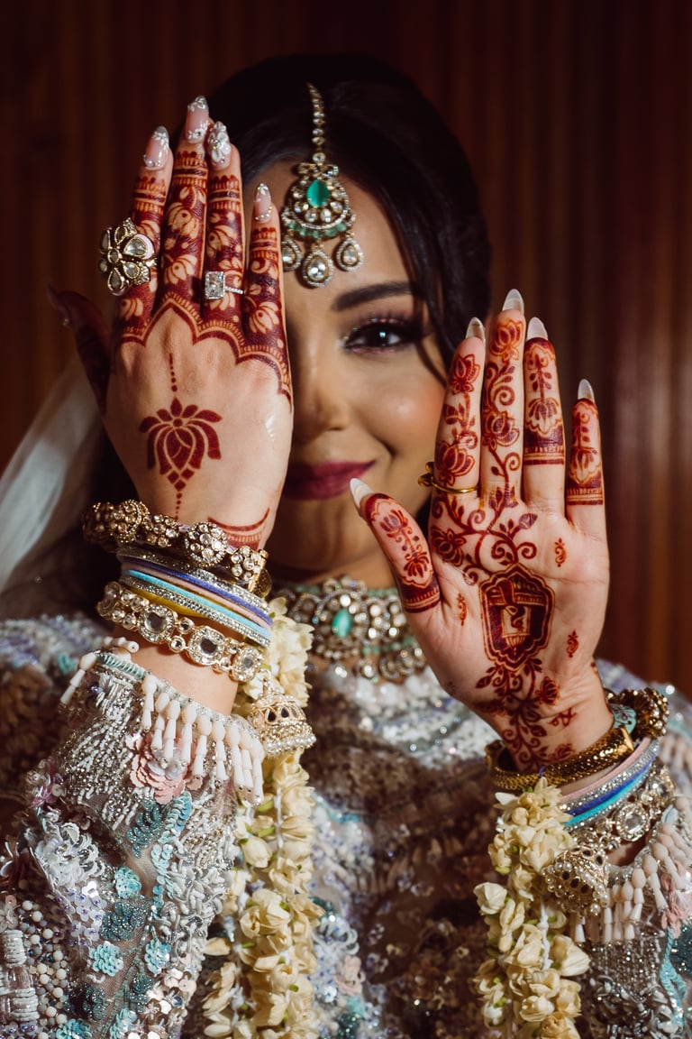Woman in traditional Indian bridal attire with henna-decorated hands, jewelry, and ornate clothing poses against a brown backdrop