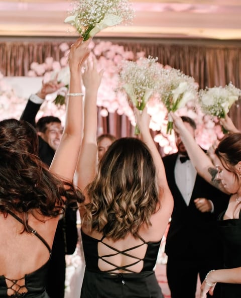 Women in black dresses catching bouquets at a wedding reception with pink and white floral decorations