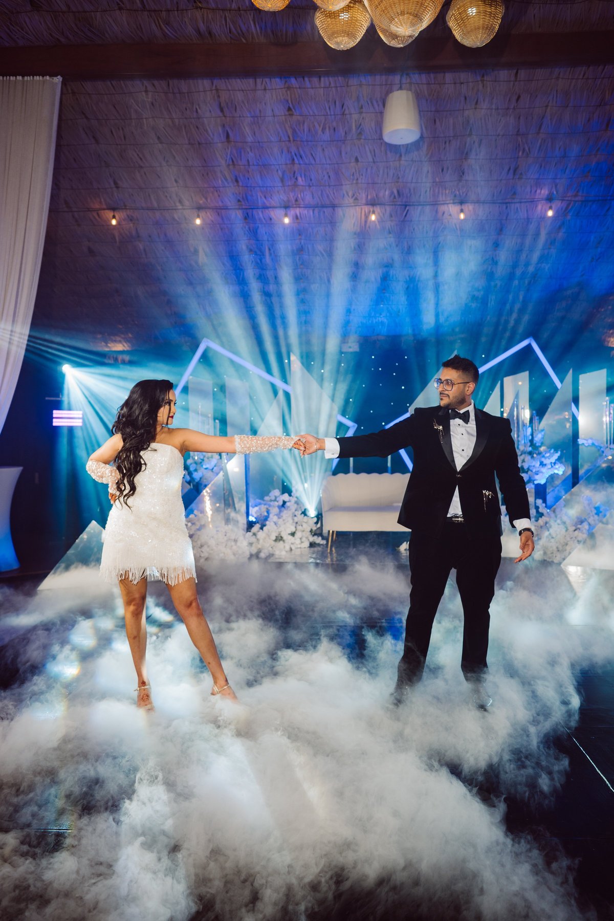 Couple dancing in white formal attire surrounded by dry ice fog with blue stage lighting and city skyline backdrop at an elegant venue
