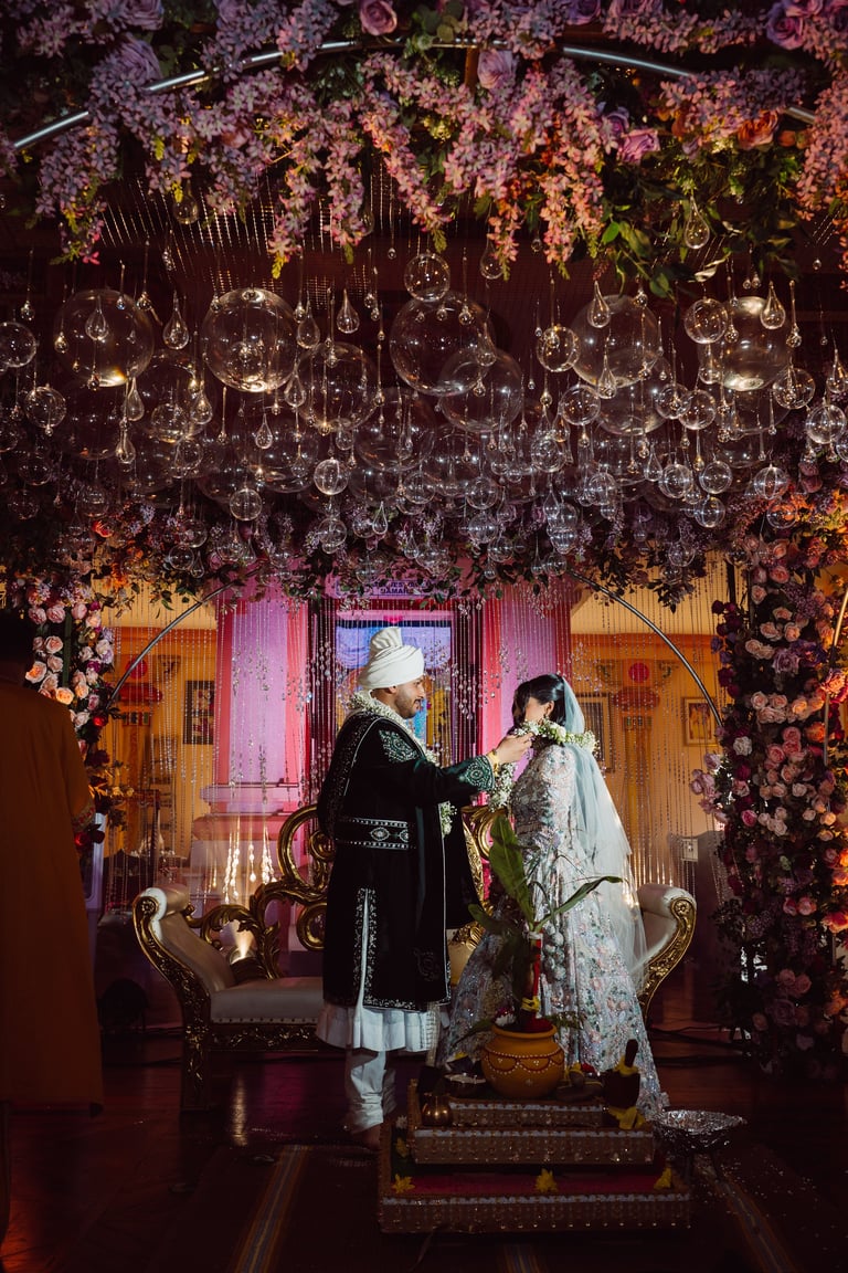 Bride and groom in traditional wedding attire standing under ornate floral arch with hanging flowers and decorative lights