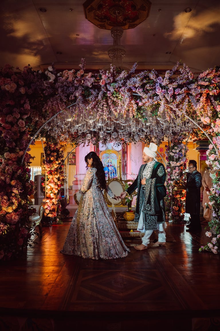 Bride in ornate lehenga walking under elaborate floral archway with guests in traditional wedding attire at Indian wedding reception.