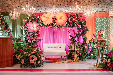 Elegant wedding stage decorated with vibrant pink and magenta flowers, lush greenery, oversize paper flowers, crystal chandeliers, and a white sofa under a pink backdrop