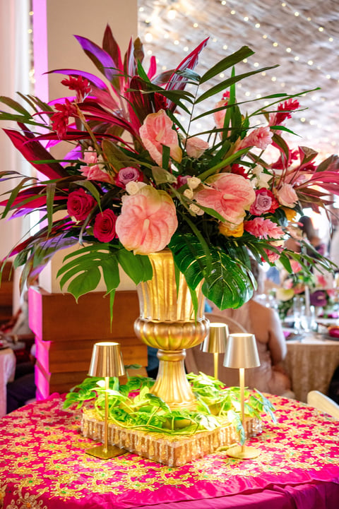Ornate gold urn overflowing with vibrant pink and red flowers and tropical foliage on a pink patterned table at an elegant event