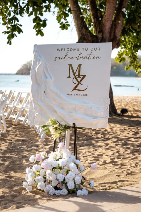 Beach wedding welcome sign with initials MZ dated May 23rd 2026, decorated with white flowers beneath a shaded tree on sandy shore