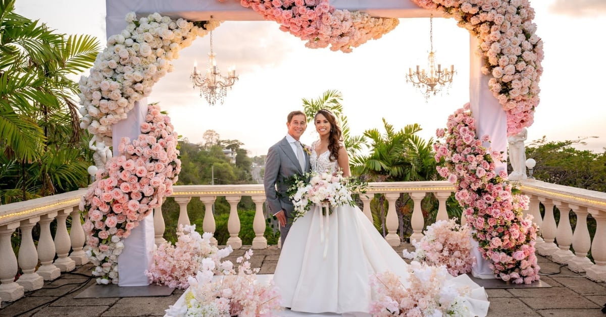 Bride and groom kiss under an ornate floral arch decorated with pink flowers and chandeliers on a stone terrace.