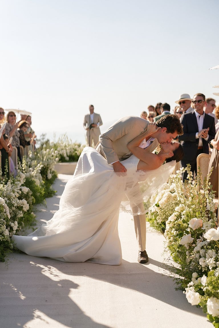 Groom dipping bride in white dress at outdoor wedding ceremony with white flower arrangements and guests in background