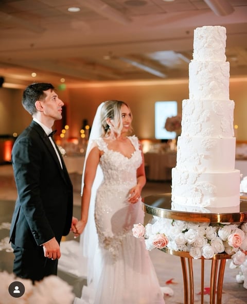 Bride and groom admiring a tall white tiered wedding cake decorated with white and pink flowers at their reception