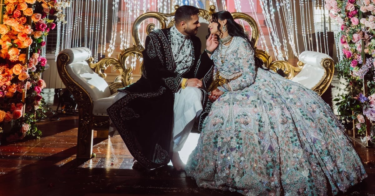 Couple in formal attire kissing on ornate gold sofa with floral decorations and elaborate backdrop