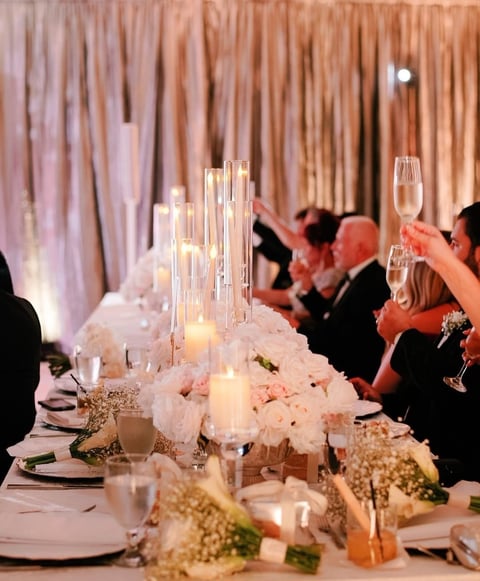 Elegant wedding reception table with white floral centerpieces, lit candles, champagne glasses, and guests toasting in the background