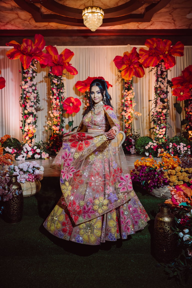 Woman in ornate colorful lehenga posing in elaborate floral setting with large red flowers and crystal chandelier
