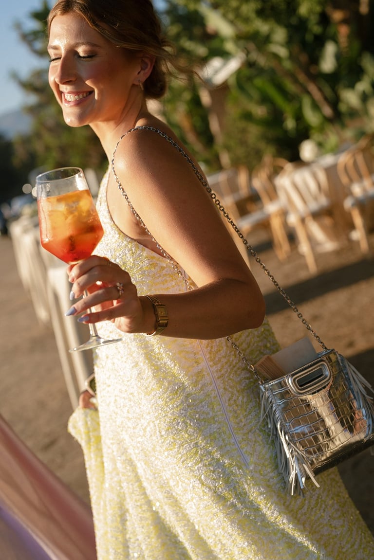Smiling woman in a yellow sequin dress holding a cocktail at an outdoor event