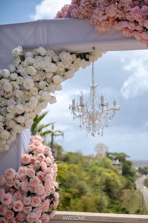 Elegant outdoor wedding ceremony setup with pink and white floral arrangements, white fabric draping, crystal chandelier, and scenic garden backdrop