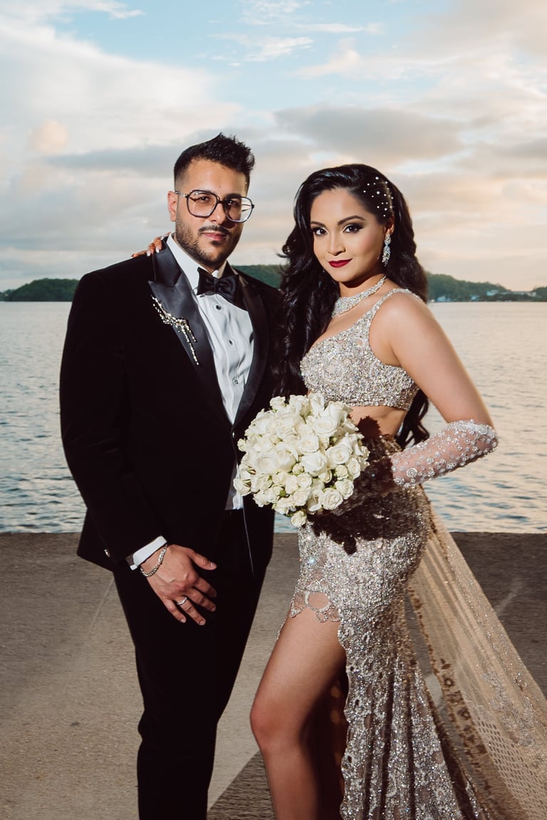 Bride and groom pose together on a beach at sunset holding white bouquet, wearing formal wedding attire