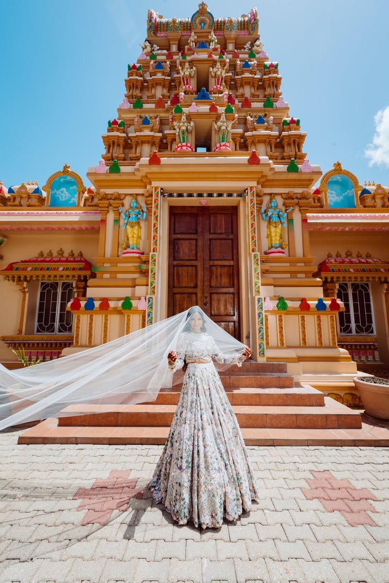 Bride in white wedding dress with flowing veil standing before ornate Hindu temple with colorful tiered gopuram and decorated entrance