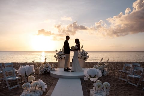 Bride and groom stand on a decorated platform for a beach wedding ceremony at sunset with white flowers lining the aisle
