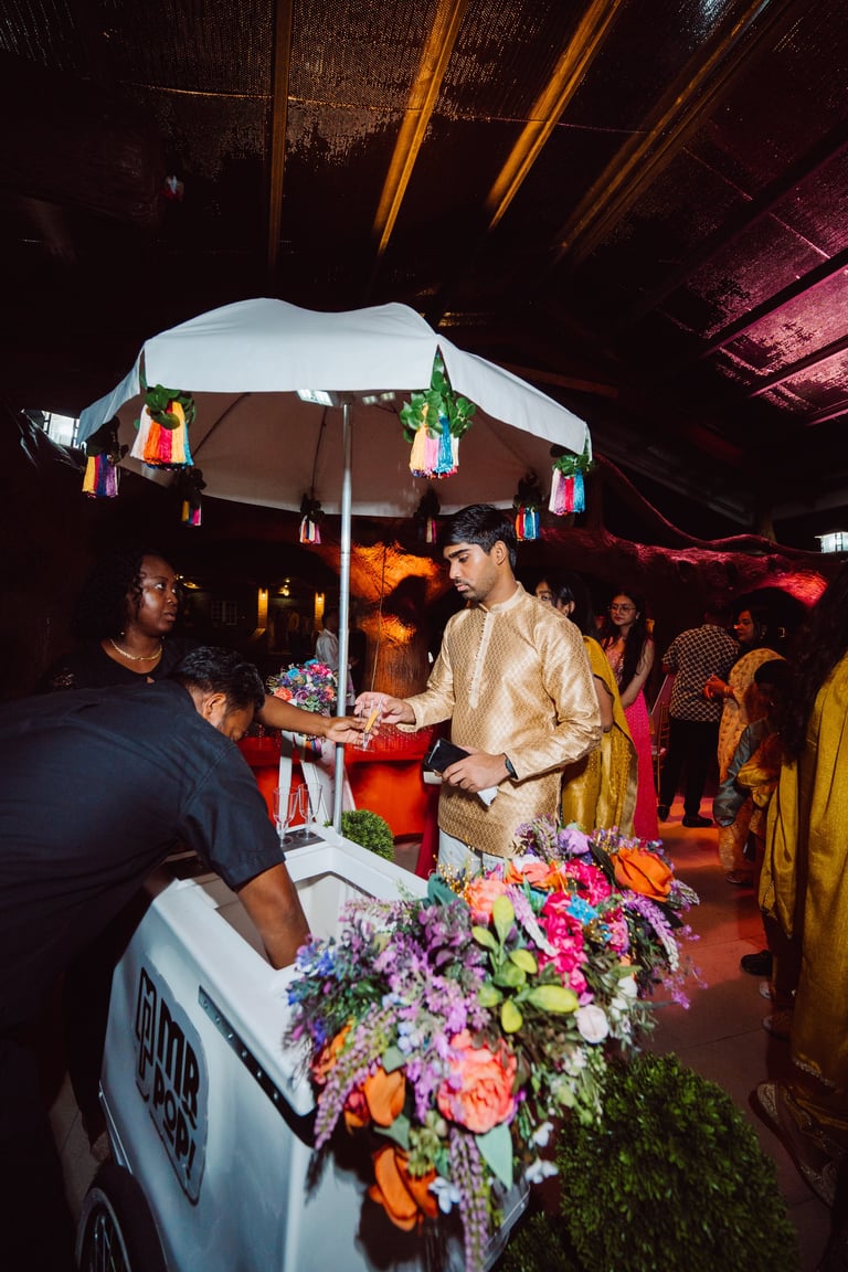 Man in beige shirt at outdoor vendor stall decorated with colorful flowers and rainbow lanterns under white umbrella at evening event
