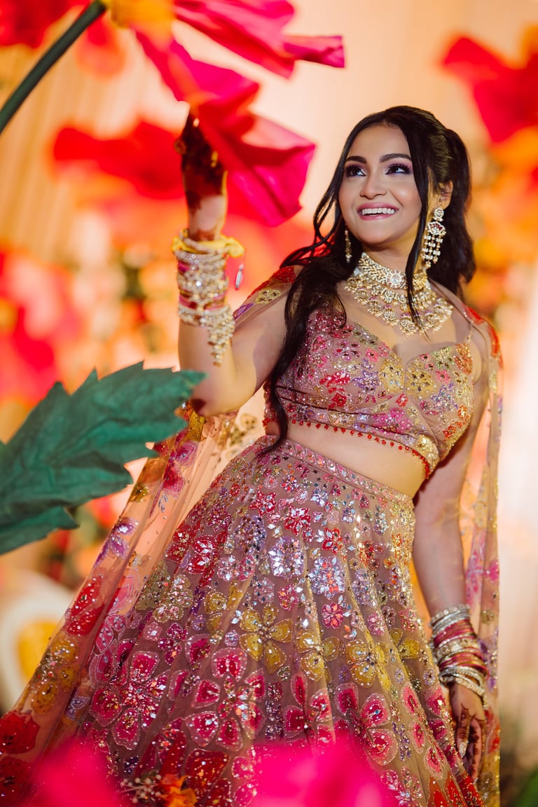 Woman in ornate pink and gold embroidered traditional Indian lehenga with jewelry against vibrant red and orange floral backdrop