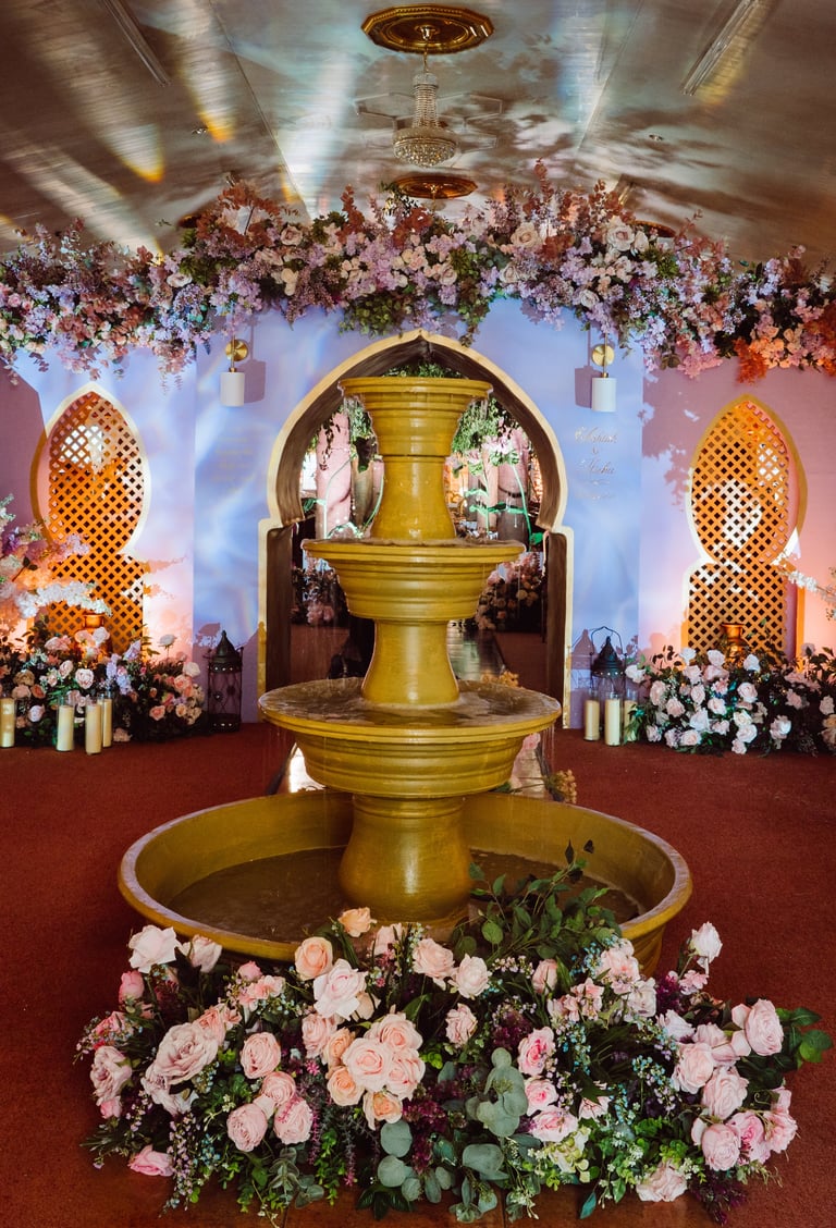 Ornate golden tiered fountain surrounded by pink and white flowers in an elegantly decorated indoor venue with floral arrangements and decorative lighting